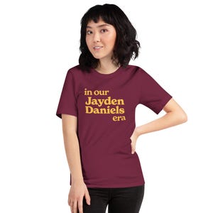 May include: A maroon t-shirt with the text "in our Jayden Daniels era" in yellow.