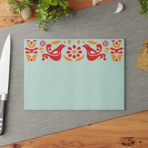 Può includere: Un tagliere di colore azzurro chiaro con un design di arte popolare rosso e arancione con uccelli e fiori. Il design è sul bordo superiore del tagliere.