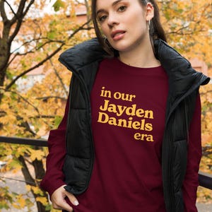 May include: A maroon long-sleeved sweatshirt with the text "in our Jayden Daniels era" printed in yellow.