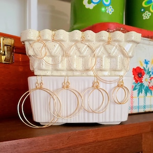 May include: A collection of gold-colored hoop earrings in various sizes. The earrings are displayed on a white, textured ceramic dish. The background includes vintage kitchenware in green and red, creating a retro aesthetic.
