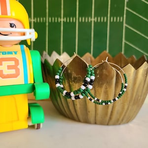 May include: A pair of hoop earrings with green, white, and black beads. The earrings are on a gold-colored surface.