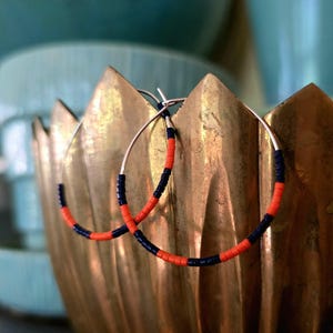 Op de afbeelding: Zilveren oorringen met afwisselende secties van oranje en marineblauwe kralen. De oorbellen worden gepresenteerd in een goudkleurige, decoratieve kom tegen een wazige turquoise achtergrond. Een eenvoudig, maar stijlvol accessoire.