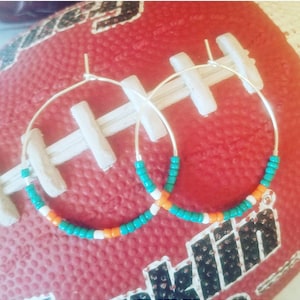 May include: A pair of silver hoop earrings with turquoise, white, and orange beads. The earrings are on a red and white football.