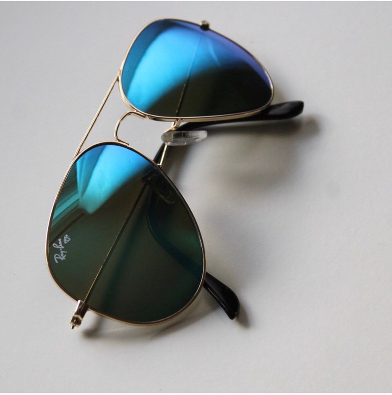 Puede incluir: Gafas de sol estilo aviador con montura dorada y lentes azules espejadas. Las lentes reflejan un tono azul vibrante, y la montura presenta un dise&ntilde;o de doble puente. Las patillas son negras, y el logotipo de Ray-Ban es visible en una lente.