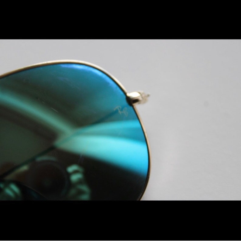 Puede incluir: Primer plano de gafas de sol de aviador con montura dorada y lentes azul-verdosas. El logotipo de Ray-Ban es visible en la lente. Las gafas de sol est&aacute;n sobre un fondo blanco.