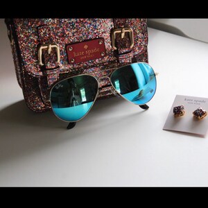 Puede incluir: Un bolso Kate Spade brillante, gafas de sol aviador con montura dorada y lentes azules, y pendientes de bot&oacute;n Kate Spade. El bolso tiene hebillas doradas y una etiqueta de cuero roja. Las gafas reflejan el entorno.