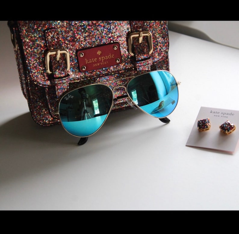 Puede incluir: Un bolso Kate Spade brillante, gafas de sol aviador con montura dorada y lentes azules, y pendientes de bot&oacute;n Kate Spade. El bolso tiene hebillas doradas y una etiqueta de cuero roja. Las gafas reflejan el entorno.