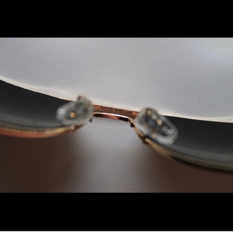 Puede incluir: Primer plano de gafas de sol Ray-Ban. La montura dorada sostiene lentes oscuras y almohadillas nasales transparentes. El texto "RAY-BAN 62014" est&aacute; impreso en la montura.