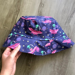May include: A blue bucket hat with a space-themed print featuring green aliens in pink flying saucers. The hat has a dark blue background with white stars and planets.