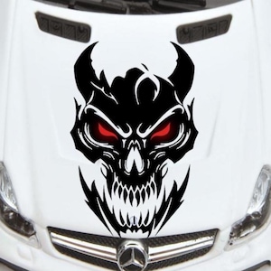 May include: A black skull decal with red glowing eyes and sharp teeth is affixed to the hood of a white car. The decal features horns and a detailed, menacing design, creating a bold statement.