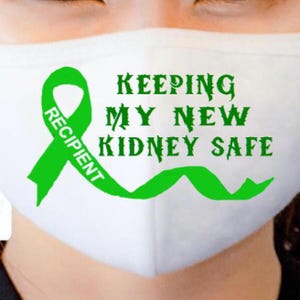 May include: White face mask with green text and a green ribbon. The text reads "KEEPING MY NEW KIDNEY SAFE" and "RECIPIENT". The ribbon is in the shape of an awareness ribbon.