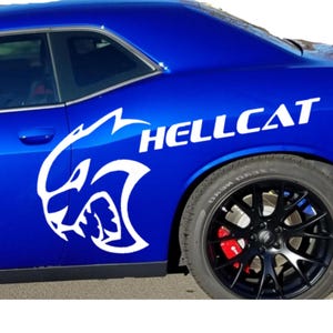 May include: A bright blue car side panel featuring a large white graphic of a stylized cat head logo and the text HELLCAT in bold white capital letters. The vehicle has a black multi-spoke wheel with a visible red brake caliper behind the spokes.