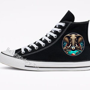 以下が含まれることがあります： 青いストライプの白いビリヤード台でビリヤードをしているゾウのグラフィックが描かれた、黒いハイカットキャンバススニーカー。ゾウはビリヤードキューを持ち、8番ボールを狙っています。「Be Legendary Too」という文字が靴の側面に書かれています。