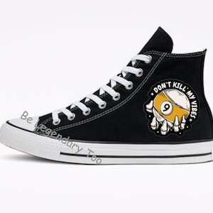 以下が含まれることがあります： 白い靴ひもが付いた黒いハイカットキャンバススニーカー。ビリヤードボールのグラフィックと「Don't Kill My Vibes」というテキストが描かれています。