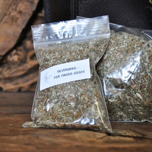 May include: Two clear plastic bags filled with dried Silverweed - Five Finger Grass. Each bag has a white label with the product name. The bags are on a wooden surface with a dark leather bag in the background.
