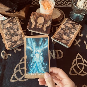 Peut inclure: Plusieurs cartes de tarot et boîtes en bois avec des illustrations mystiques, disposées sur un tissu noir avec des symboles de nœuds celtiques. Une carte présente une illustration bleue d'une figure avec le texte "Penelope Dreamweaver". Une bougie allumée et des cristaux sont également présents.