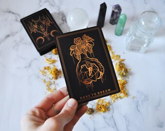May include: Black tarot card with gold foil illustration of a hand holding a geometric shape, with the words "DARE TO DREAM". A deck of cards is in the background, along with crystals and a glass bottle.