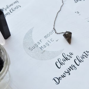 May include: A collection of spiritual items including a pendulum, crystals, and a small glass jar with incense. The words "Sugar moon Magic" and "Chakra Dowsing Charts" are visible. The pendulum hangs over a page with text.
