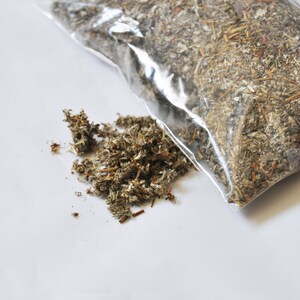 May include: A clear plastic bag filled with dried, chopped herbs, and a small pile of the herbs next to the bag. The herbs are a mix of green, brown, and tan colors, with a slightly textured appearance. The background is white.