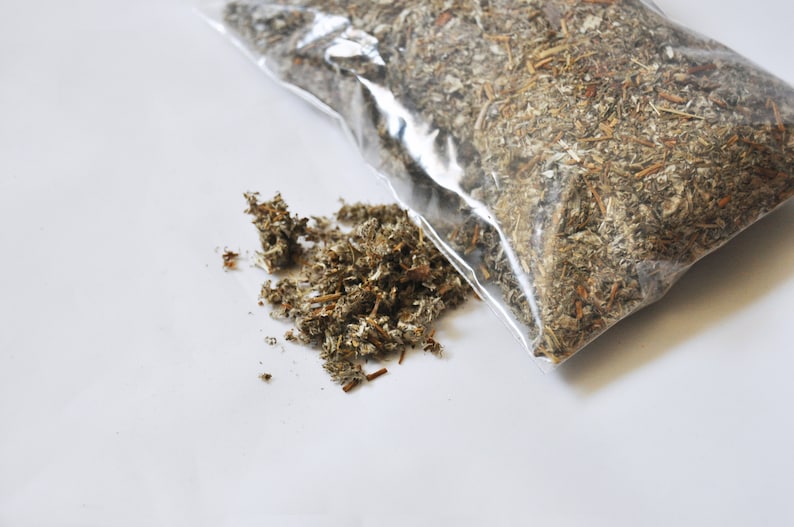 May include: A clear plastic bag filled with dried, chopped herbs, and a small pile of the herbs next to the bag. The herbs are a mix of green, brown, and tan colors, with a slightly textured appearance. The background is white.