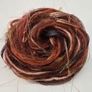 May include: A circular decorative accessory crafted from a dense, spiraling arrangement of textured fibers and ribbons in shades of deep copper, bronze, dark brown, and rust, featuring loose, wispy strands extending from the central coiled form.
