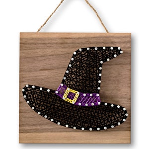 May include: A black witch hat with a purple and yellow band is strung with yarn on a wooden plaque. The plaque has a rope hanger.