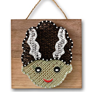 May include: A string art wall hanging featuring a green monster face with black hair and white stitches. The monster has a red smile and black eyes. The art is on a wooden background with a rope hanger.