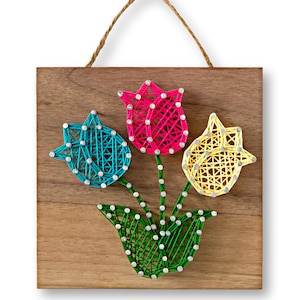 May include: A wooden wall hanging with three colorful string art tulips. The tulips are pink, blue, and yellow. The tulips are arranged in a bouquet with green leaves.