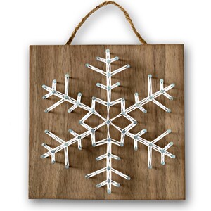 May include: A white string art snowflake design on a brown wooden background. The snowflake is created by wrapping white string around nails hammered into the wood.