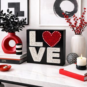 Może przedstawiać: Czarny drewniany znak z napisem "LOVE" w białych literach z nici i czerwonym sercem. Znak jest ozdobiony czerwonym sercem i białymi kropkami. Znak znajduje się na białej powierzchni z innymi elementami dekoracyjnymi.