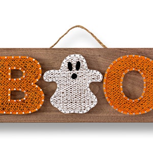 Puede incluir: Un letrero de madera con la palabra "BOO" escrita en hilo naranja. Un fantasma de hilo blanco está entre la "B" y la "O".