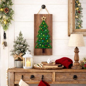 May include: A wooden Christmas tree wall hanging with a green tree design, adorned with colorful lights and a gold star. The tree is mounted on a rectangular wooden board. The scene includes a small Christmas tree, a lamp, and a decorative table.