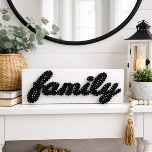 May include: A white wooden sign with the word "family" spelled out in black string art with white polka dots. The sign is displayed on a white surface, with decorative items in the background.
