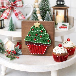 Può includere: Decorazione natalizia con un albero di Natale in filo di legno su un blocco di legno. L'albero è verde con dettagli rossi e bianchi. Vengono presentati due cupcakes con glassa bianca e ciliegie.