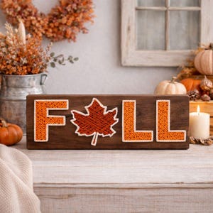 May include: Wooden sign with the word "FALL" spelled out in orange thread, with a maple leaf in place of the "A". The sign is on a wooden surface, with pumpkins and a lit candle in the background. The sign is a fall-themed decoration.