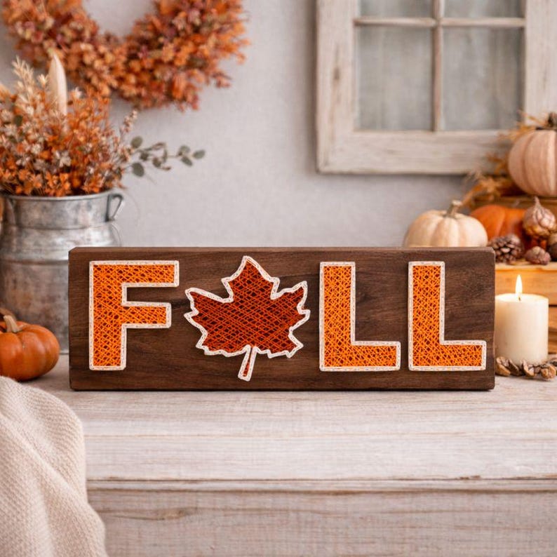 May include: Wooden sign with the word "FALL" spelled out in orange thread, with a maple leaf in place of the "A". The sign is on a wooden surface, with pumpkins and a lit candle in the background. The sign is a fall-themed decoration.