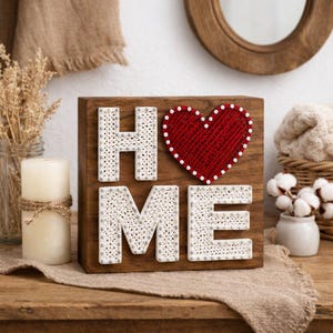 May include: Wooden sign with the word "HOME" spelled out in white string art letters and a red heart. The sign is on a wooden surface, with a candle and other decorative items in the background.