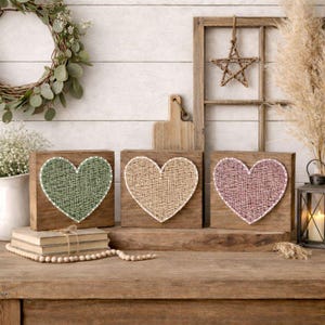 Puede incluir: Tres bloques de madera con corazones de hilo en verde, beige y rosa. Los corazones están centrados en los bloques, con un borde blanco. Los bloques se exhiben sobre una mesa de madera, con una corona y otros elementos decorativos en el fondo.