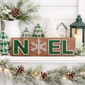 Peut inclure: Panneau en bois avec le mot "NOEL" en vert et un flocon de neige blanc. La sc&egrave;ne est orn&eacute;e de d&eacute;corations de No&euml;l, notamment des pommes de pin, de la verdure et de petits arbres d&eacute;coratifs. Une bougie allum&eacute;e est dans une lanterne verte.
