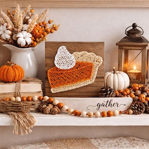 May include: A seasonal display with a string art pie slice and whipped cream on a wooden board. The display includes a white pumpkin, a lantern with a lit candle, and a vase of autumn foliage. A wooden sign says "gather".