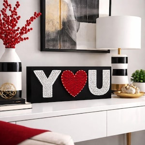 Peut inclure: Panneau rectangulaire noir avec le mot "YOU" en fil blanc et un cœur rouge. Sur une surface blanche, avec une lampe et un vase rayé noir et blanc en arrière-plan. Décoration murale.