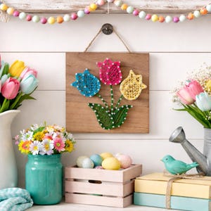 May include: A wooden wall hanging featuring string art tulips in blue, pink, and yellow. The tulips are arranged in a green vase shape. The background includes a white wall, flowers in vases, and decorative Easter eggs.
