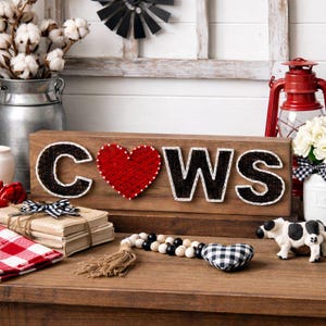 May include: Wooden sign with the word "COWS" spelled out in black letters with white stitching. The "O" is replaced with a red heart. The sign is on a wooden surface with decorative items like a cow figurine and a red lantern.