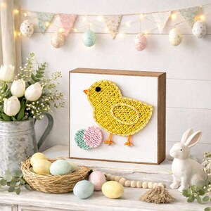Op de afbeelding: Een houten paasbord met een gele draadkunstkuiken en kleurrijke draadkunsteieren. De scène omvat een wit keramisch konijn, een eierkorf en een bloemenslinger. Lente decoratie.