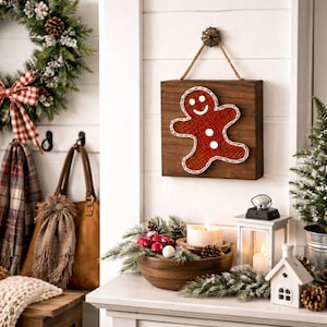Puede incluir: Decoración navideña de madera con diseño de hombre de jengibre. El hombre de jengibre rojo está cosido sobre un cuadrado de madera marrón oscuro, con un borde blanco y botones blancos. Cuelga de una cuerda de yute.