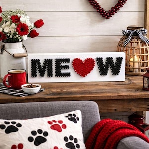 Puede incluir: Un letrero de madera blanco con la palabra "MEOW" escrita en letras negras con puntos blancos, y un corazón rojo en lugar de la "O". El letrero está sobre una mesa de madera con una taza roja, flores y un cojín con huellas de patas de gato.