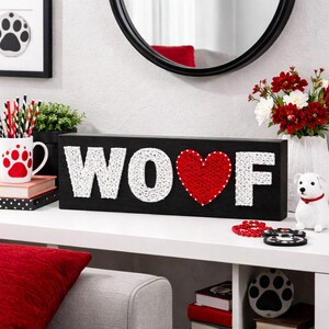 May include: A black wooden sign with the word "WOOF" in white string art, with a red heart replacing the "O". The sign is decorated with a red heart and white dots. Other items include a dog paw print mug and a small white dog figurine.