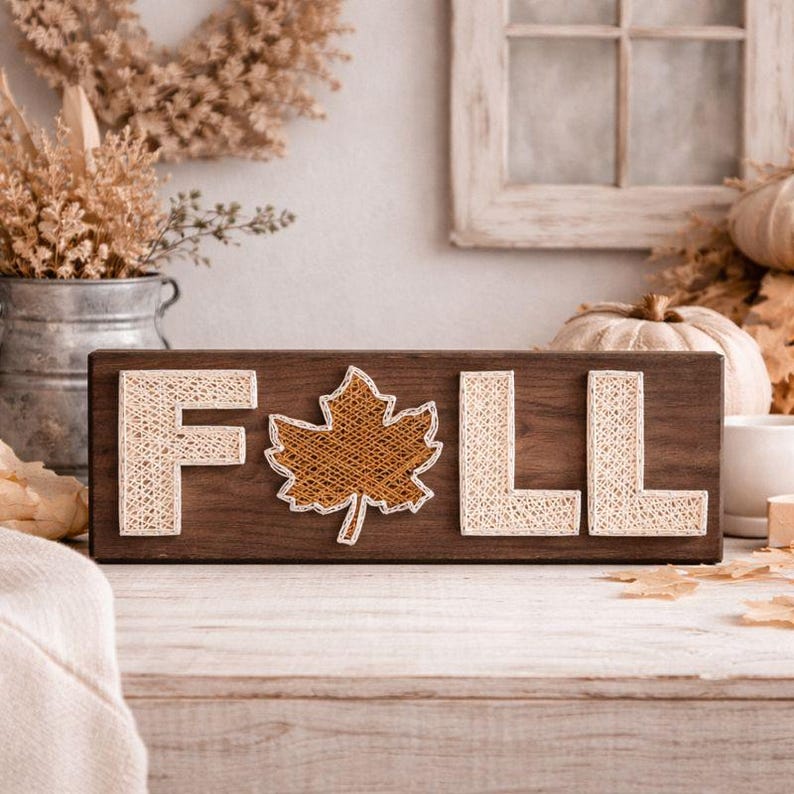 May include: A wooden sign with the word "FALL" spelled out in string art. The letters are cream-colored, and the leaf is brown. The sign is on a wooden surface, with fall-themed decorations in the background.