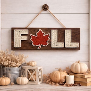Könnte beinhalten: Ein Holzschild mit dem Wort "FALL" in String-Art. Das "F" und "LL" sind cremefarben, das "A" ist ein rotes Ahornblatt. Das Schild hängt über einer Ausstellung von Kürbissen, Trockenblumen und einer Kerze.