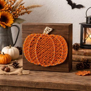 May include: A wooden block with an orange string art pumpkin design. The pumpkin is made of orange thread and silver-colored pins. The stem is a light beige color. The background includes a bat, a lantern, and other fall decorations.